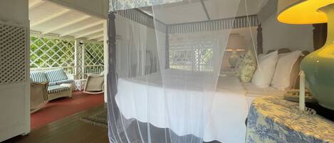 A bedroom with a four-poster bed, a canopy, a nightstand, a lamp, and a view of the outdoors.
