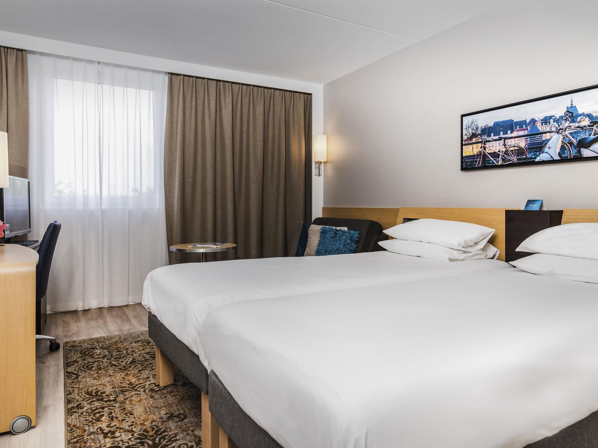 A hotel room with a bed, a desk, a chair, a small table, and a painting on the wall.