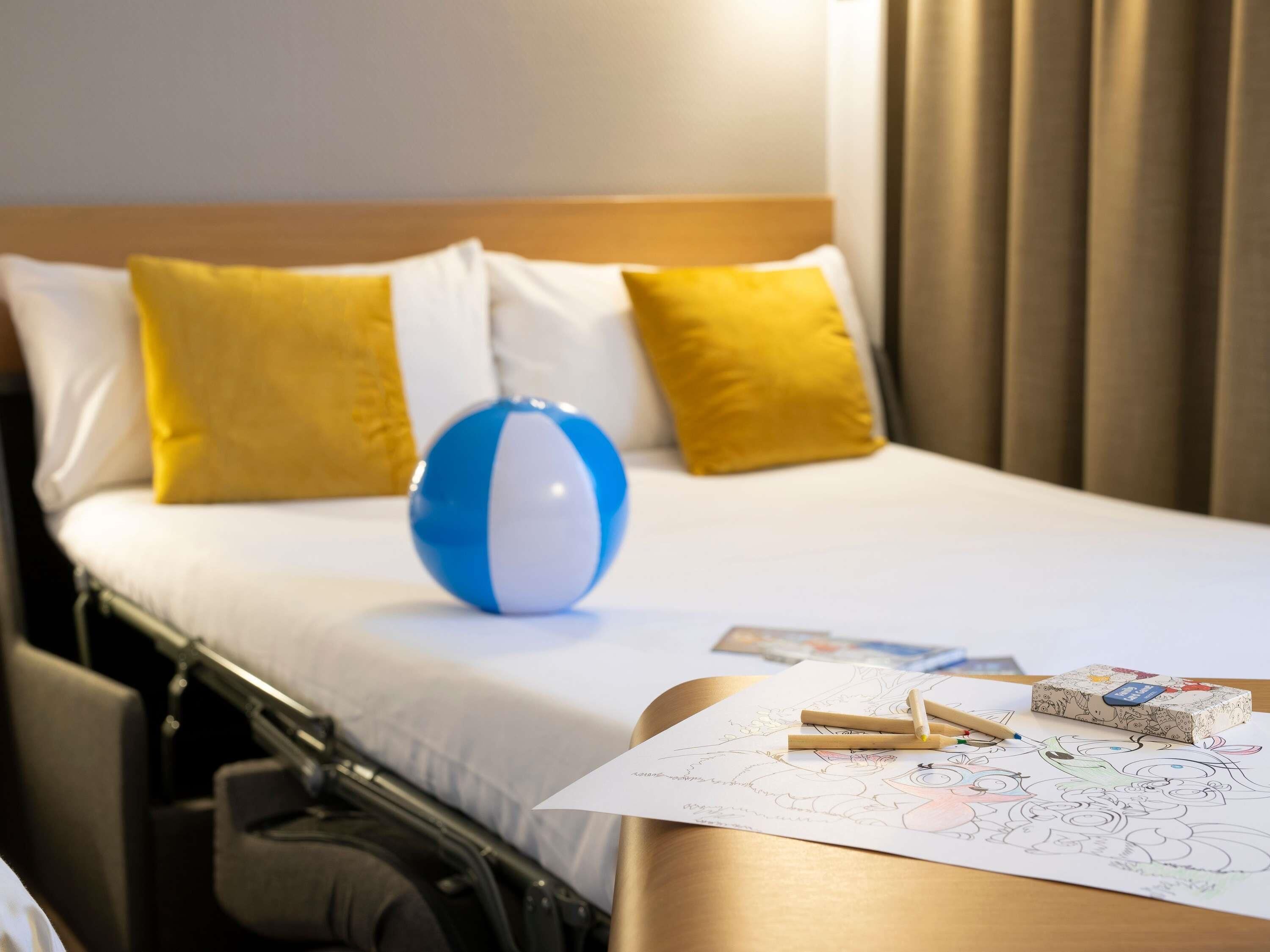 A hotel room with a bed, a blue and white beach ball, yellow pillows, a bedside table with a coloring book and pencils.