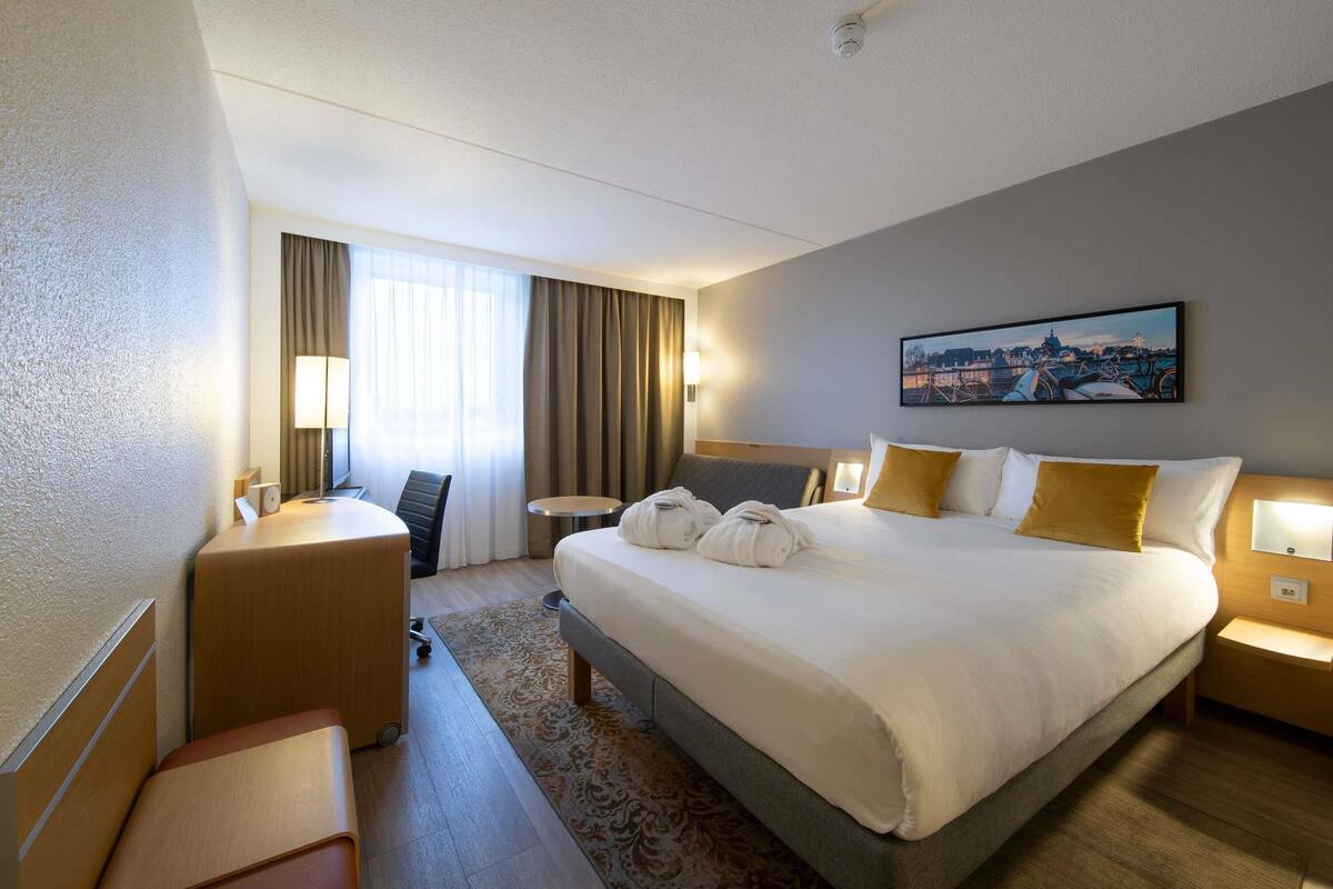 A hotel room with a bed, bedside table, desk, chair, and a painting on the wall.