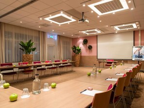 Un salón de conferencias con largas mesas, sillas, una pantalla de proyector y plantas en macetas.