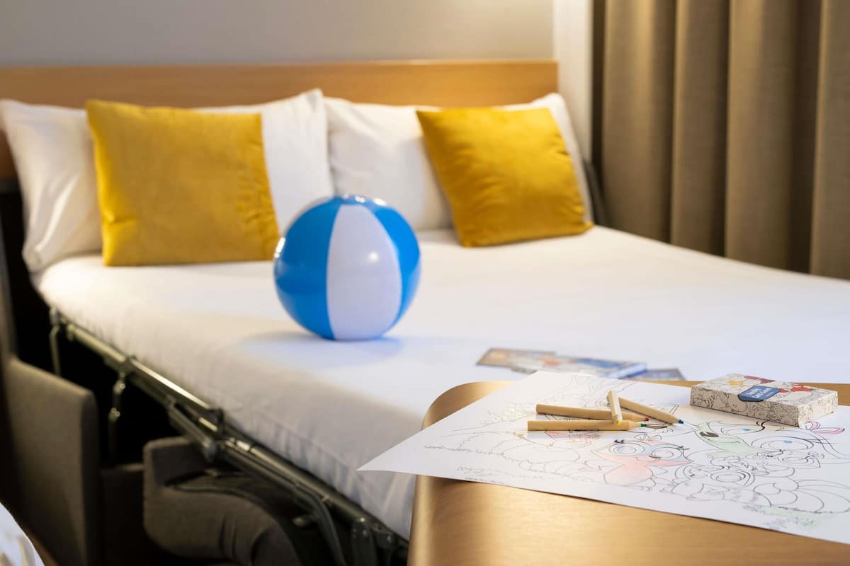 Ein Hotelzimmer mit einem Bett, einem blau-weißen Strandball, gelben Kissen und einem Nachttisch mit einem Malbuch und Bleistiften.