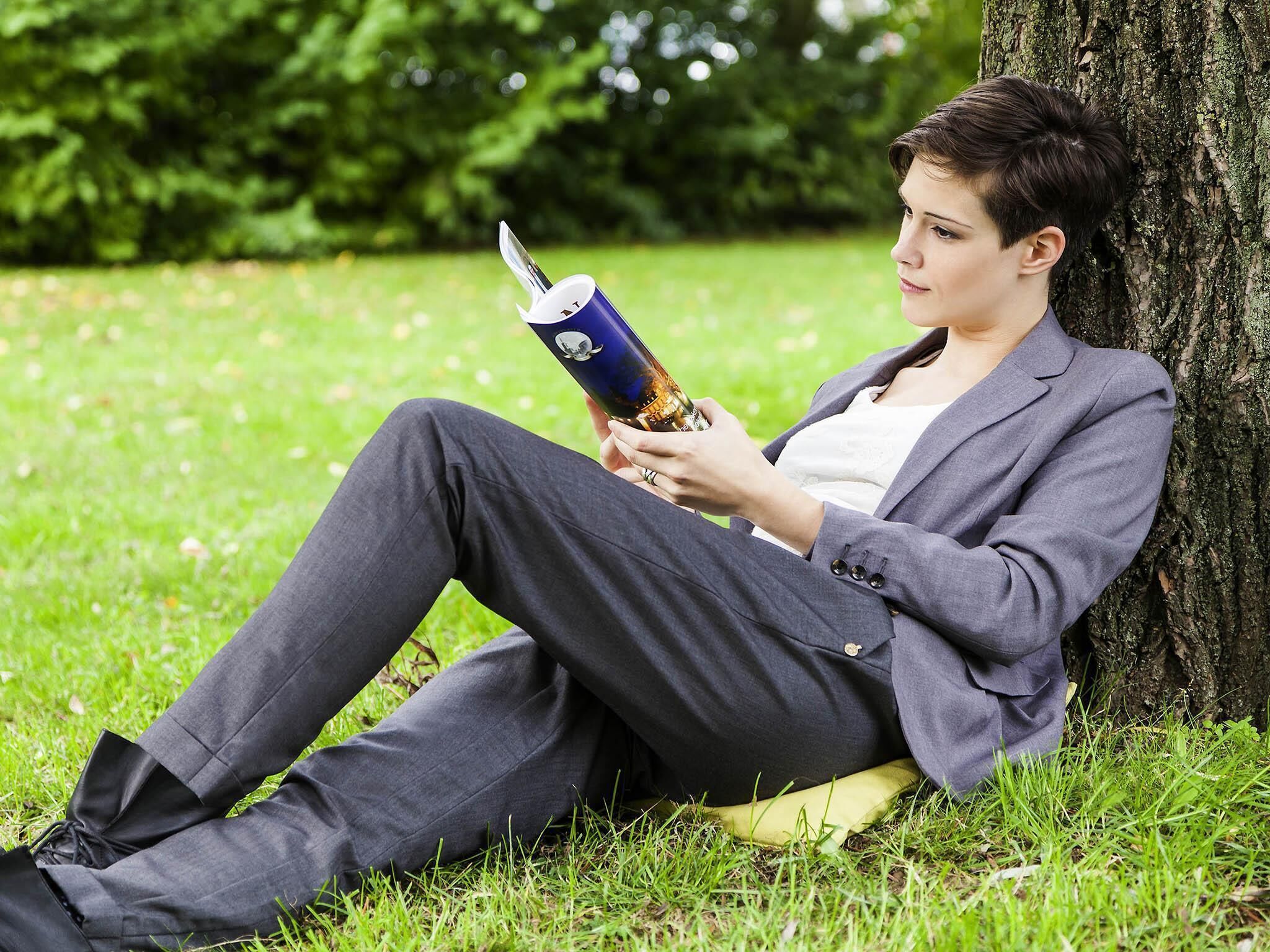 Une personne assise sur l’herbe, près d’un arbre, est en train de lire un livre.