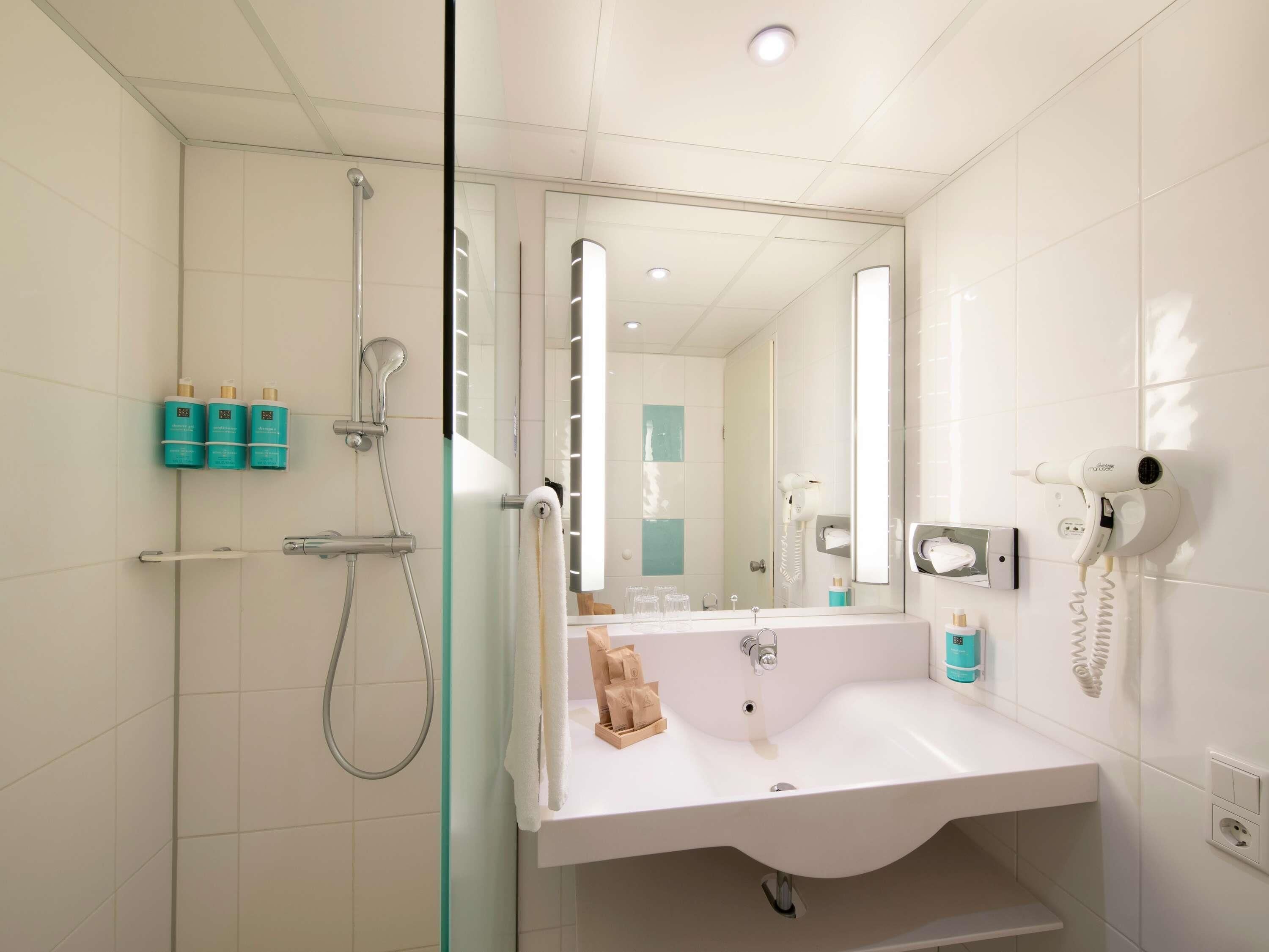 Un baño moderno con ducha envuelta en vidrio, un espejo grande, un lavamanos blanco y un secador de pelo montado en la pared.