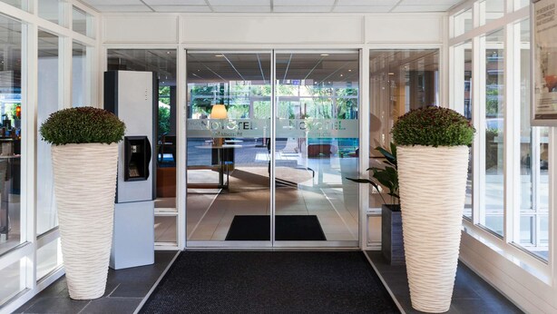 A hotel entrance with a glass door, a black mat, and two large potted plants. - Novotel Maastricht (Maastricht)