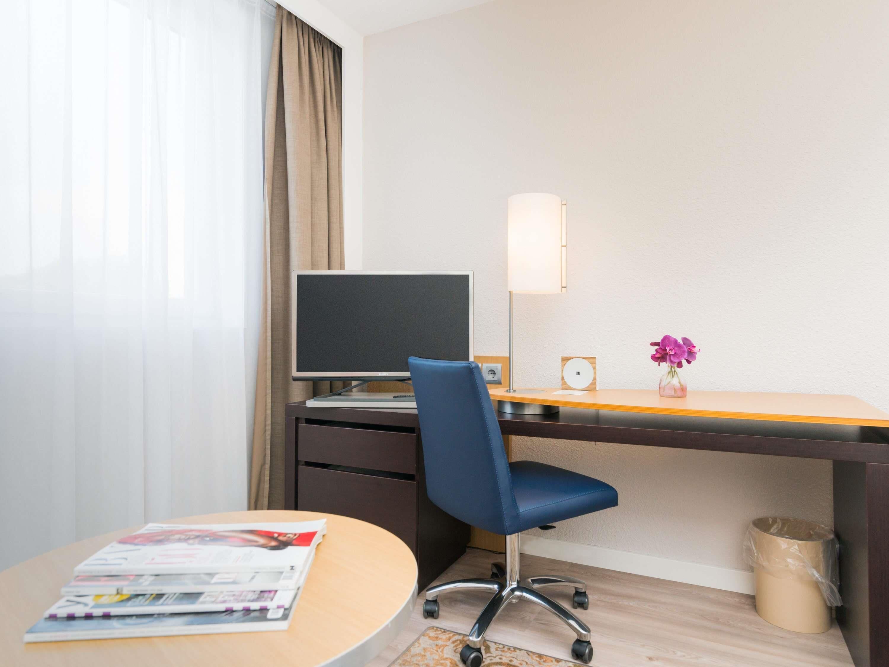 Ein Hotelzimmer mit Schreibtisch, Stuhl, Computer, Lampe, Mülleimer und einem kleinen Tisch mit Zeitschriften.