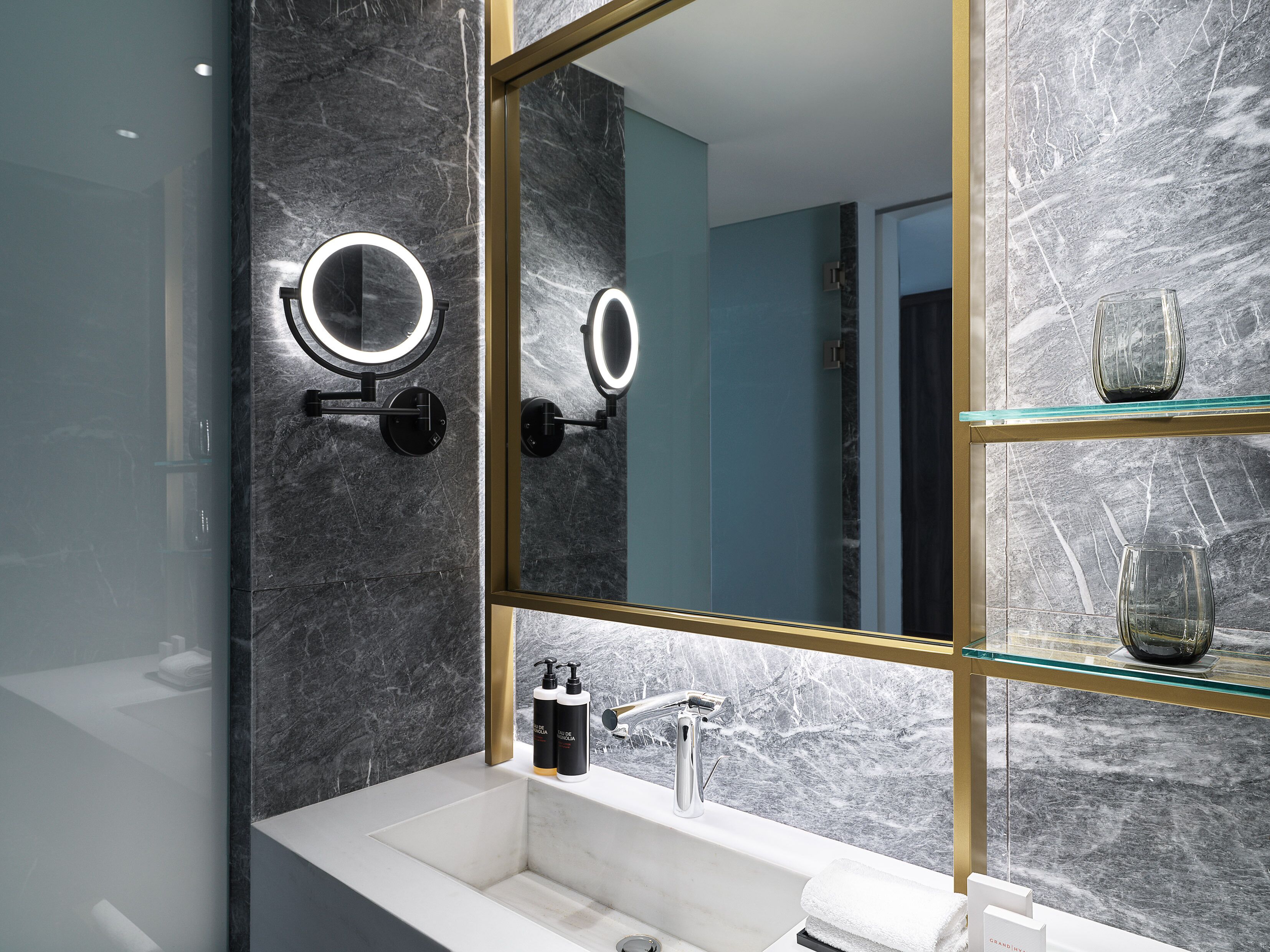 A modern bathroom with a large mirror, two wall-mounted lights, a glass shelf with decorative items, and a white sink.