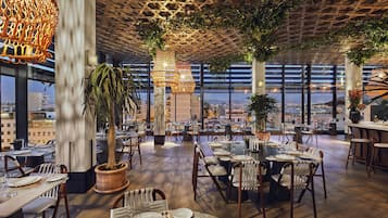 A restaurant with wooden flooring, tables set for dining, hanging wicker chandeliers, and large windows offering a city view.