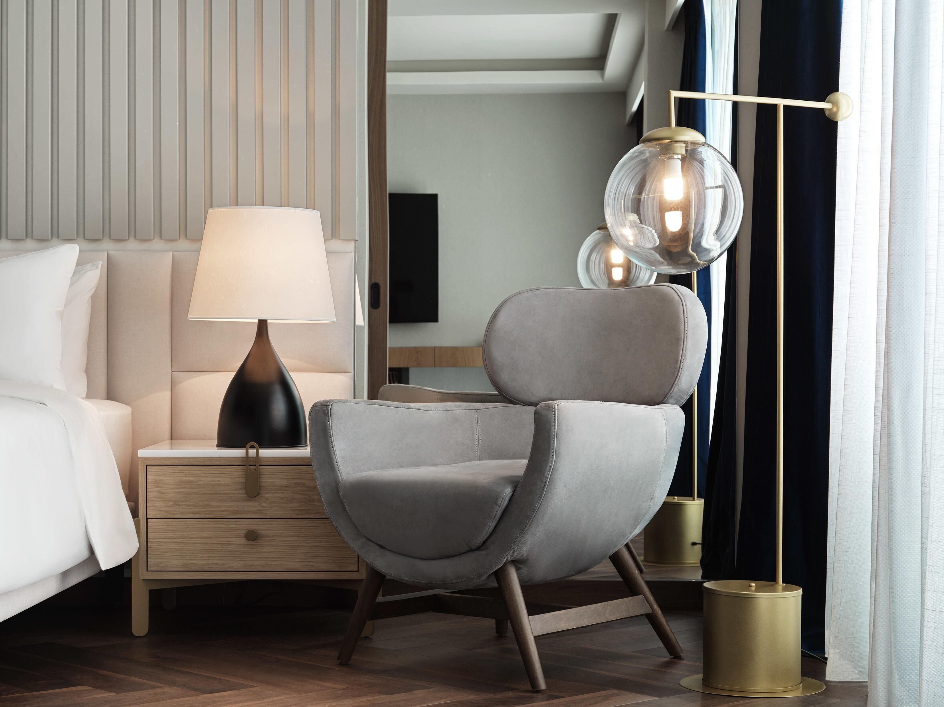 A modern hotel room with a bed, a grey armchair, a bedside table with a lamp, and a floor lamp.