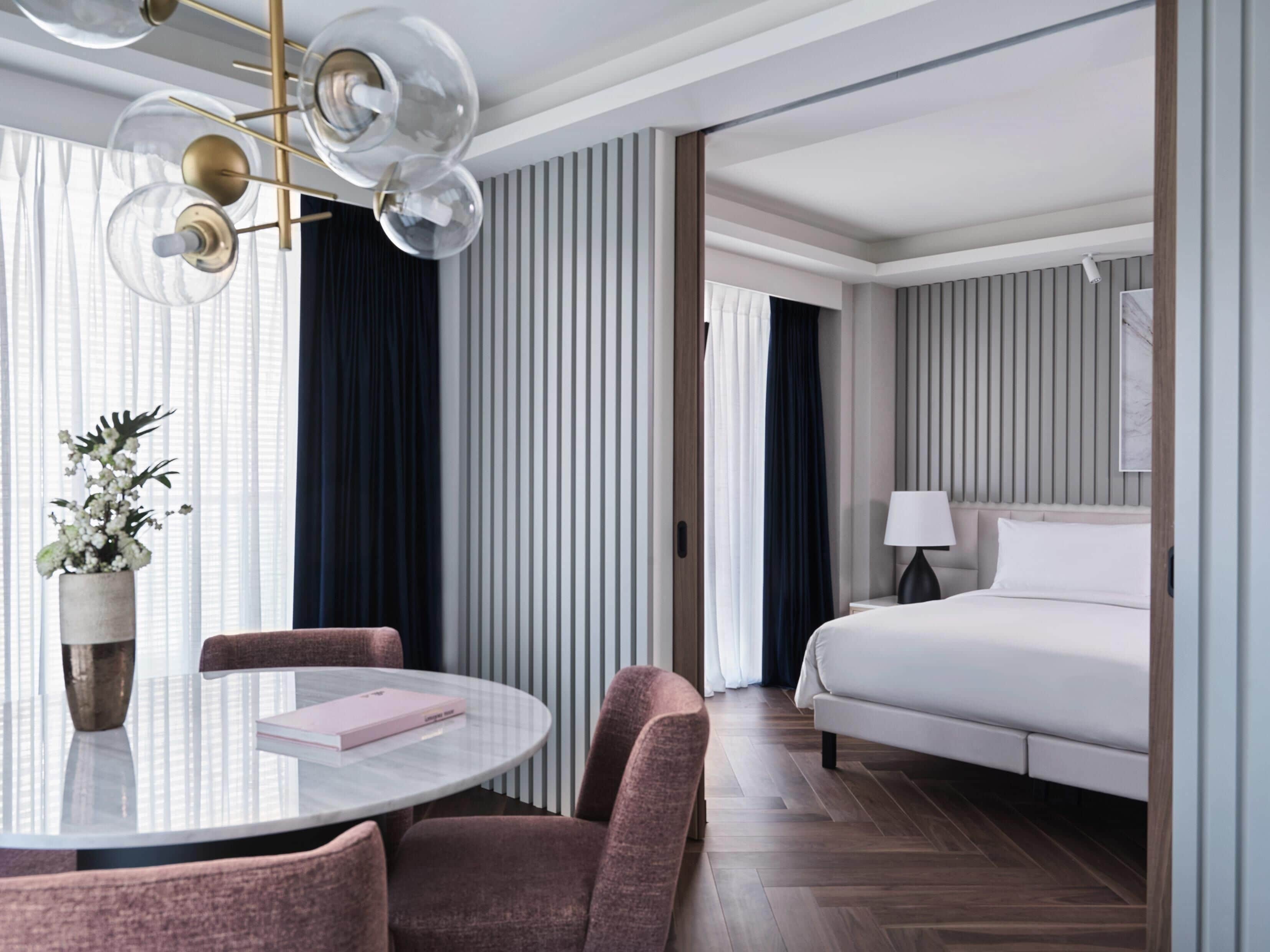 A modern hotel room with a large bed, a round table with chairs, a potted plant, and a ceiling light fixture.