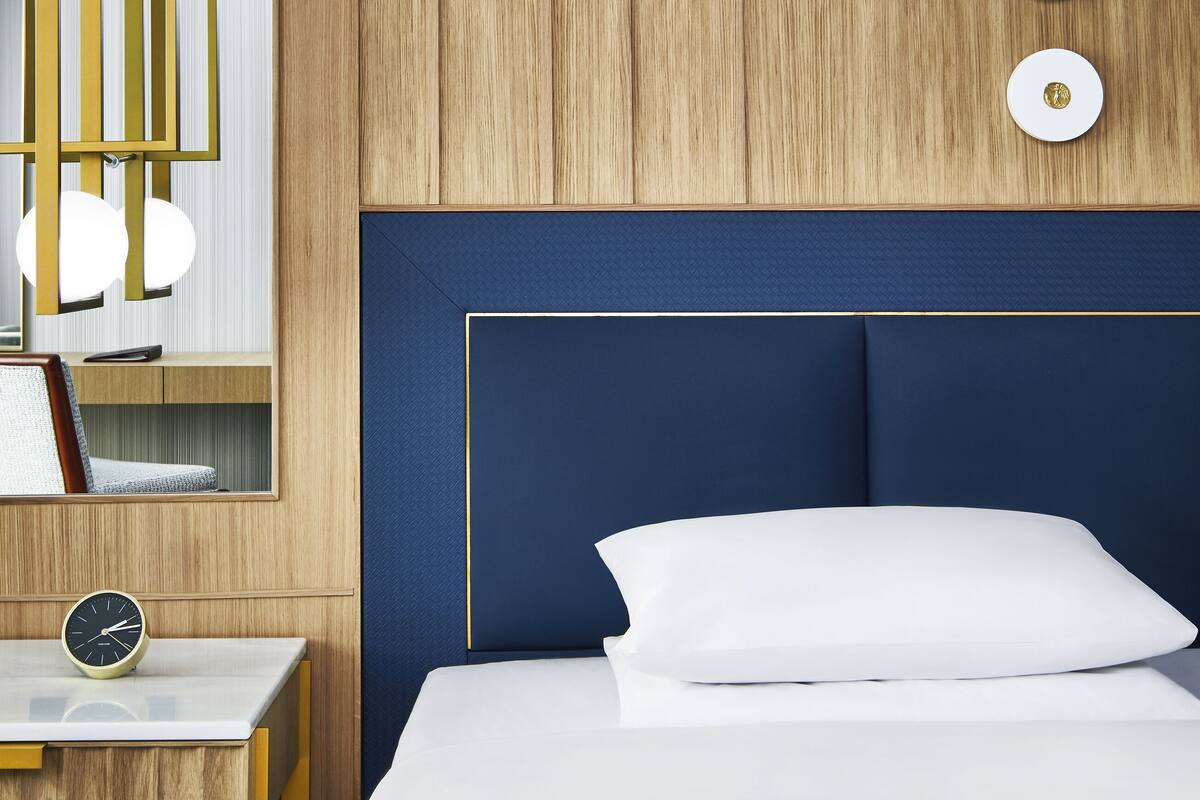 Une chambre avec un mur lambrissé en bois, une tête de lit bleue, une table de chevet avec une horloge et un tableau encadré.