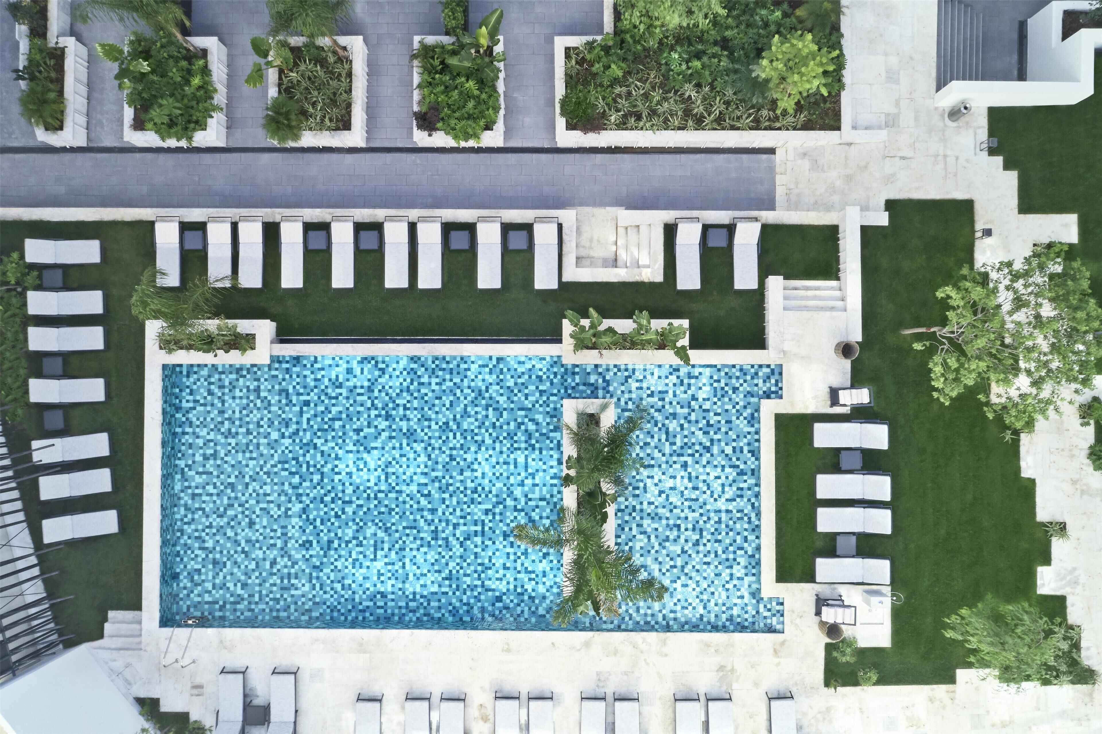 An aerial view of a modern hotel with a rectangular swimming pool surrounded by lounge chairs and greenery.