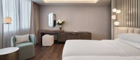 A modern hotel room with a large bed, a seating area with a sofa and armchair, a vanity with a mirror and flowers, and a bedside table.