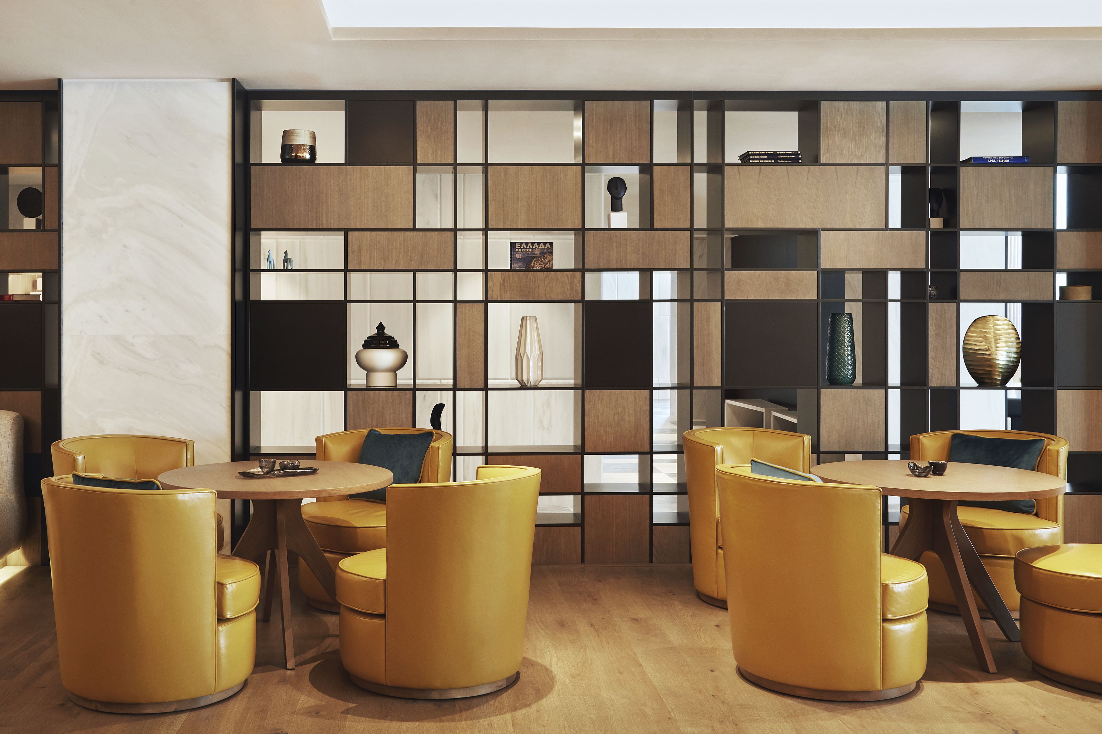 Un intérieur moderne avec des tables rondes et des chaises jaunes, un plancher en bois et un mur orné d’une grille d’étagères contenant divers objets.