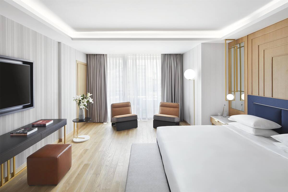 Une chambre d’hôtel moderne dotée d’un grand lit, d’une télévision à écran plat, d’un bureau avec une chaise et d’un petit coin salon avec deux fauteuils.