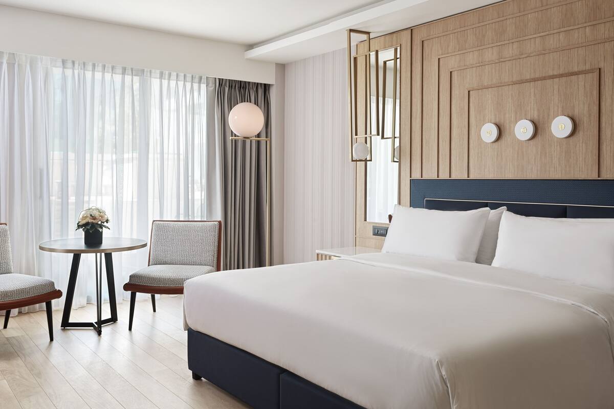 Une chambre d’hôtel moderne avec un grand lit, deux fauteuils, une petite table avec un vase et une grande fenêtre avec des rideaux.
