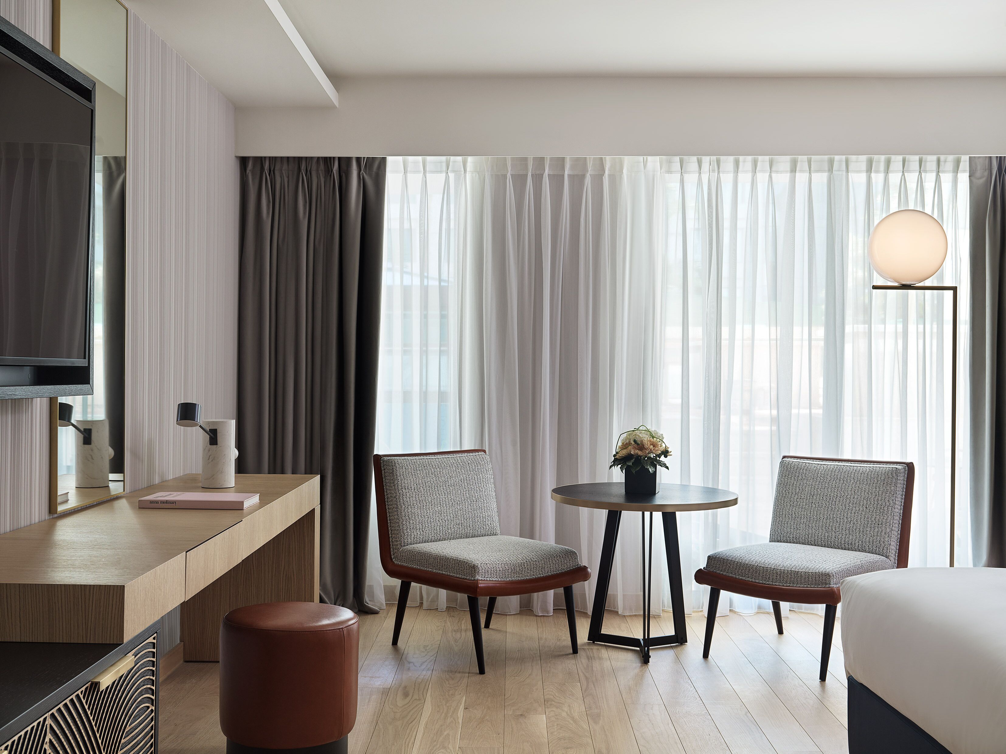 A hotel room with a bed, a desk with a mirror, two chairs, a small table with a vase, and a floor lamp.