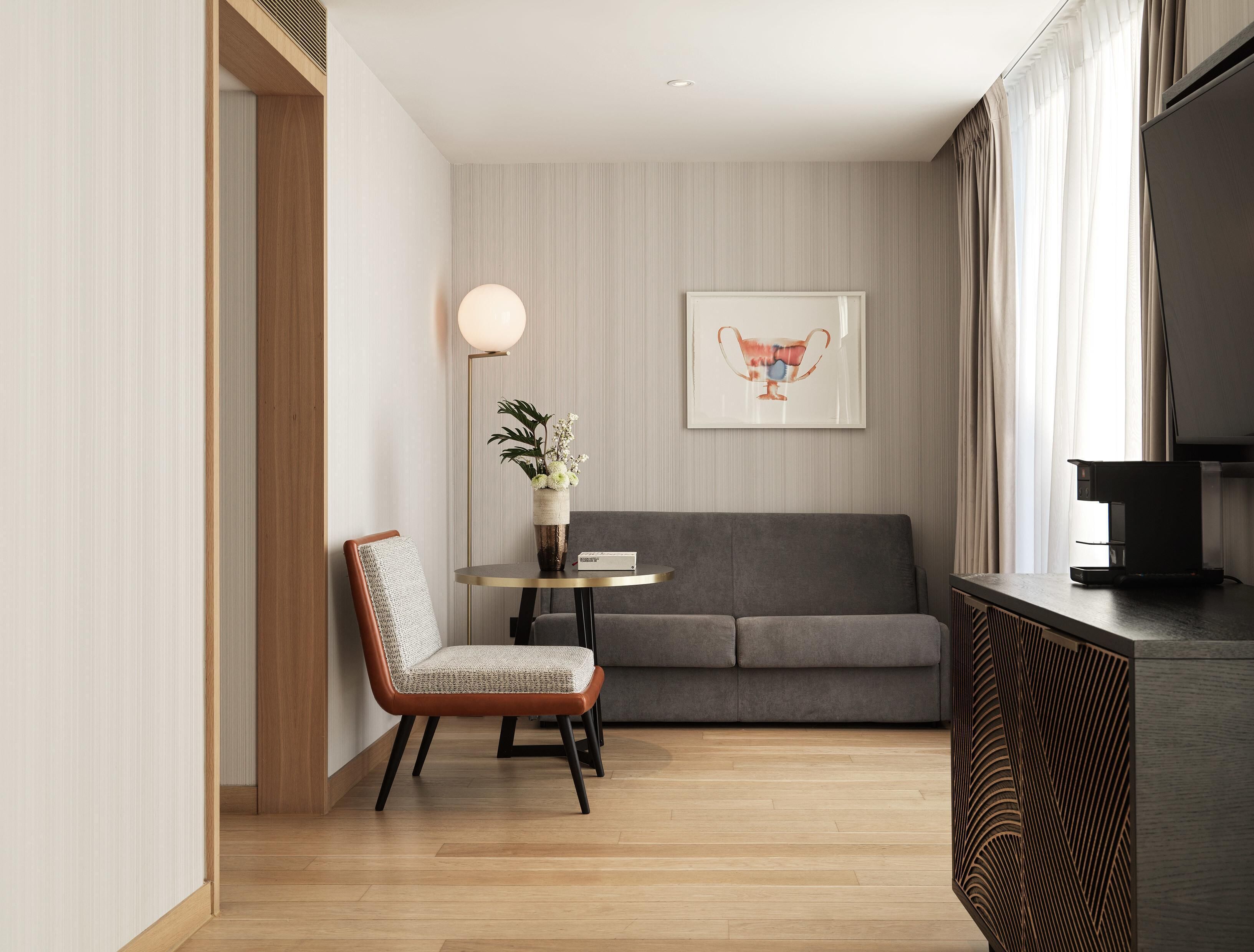 A modern living room with a grey sofa, a wooden floor, a round table with a vase of flowers, and a framed picture on the wall.