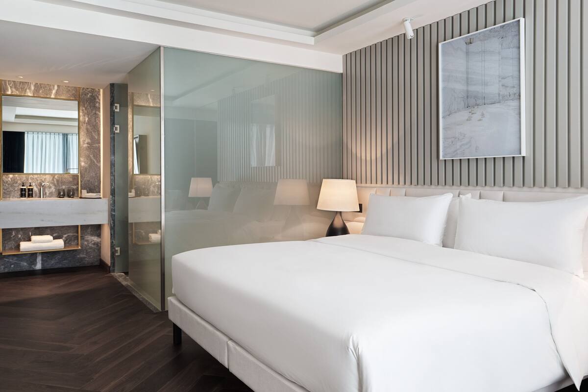 Une chambre d’hôtel moderne dotée d’un grand lit, d’une salle de bain entièrement vitrée et d’un meuble-lavabo en marbre.