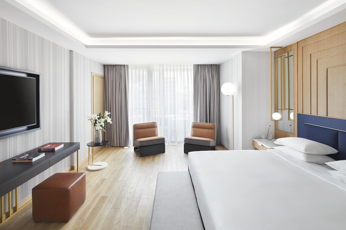 A modern hotel room with a large bed, a flat-screen TV, a desk with a chair, and a small seating area with two chairs.