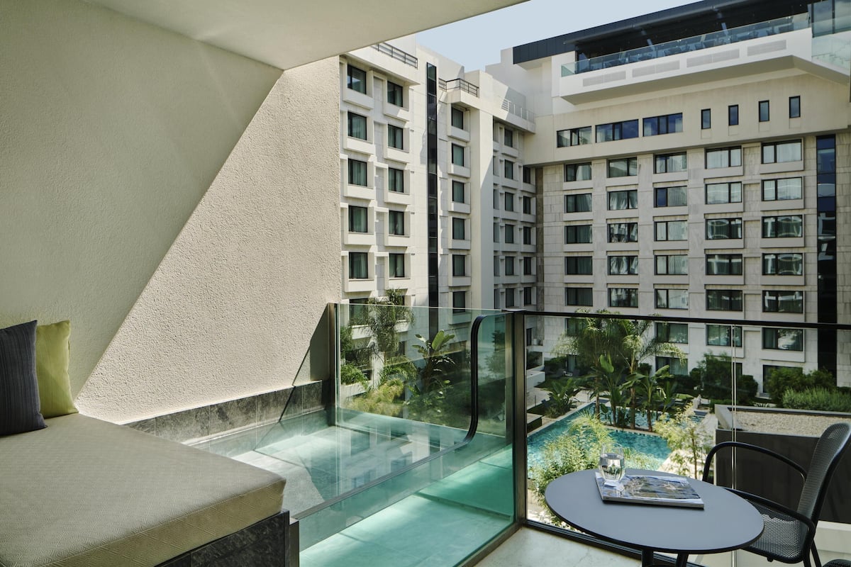 Un balcon moderne d’hôtel, doté d’une rambarde en verre, d’un espace salon et offrant une vue sur une piscine et les bâtiments environnants.