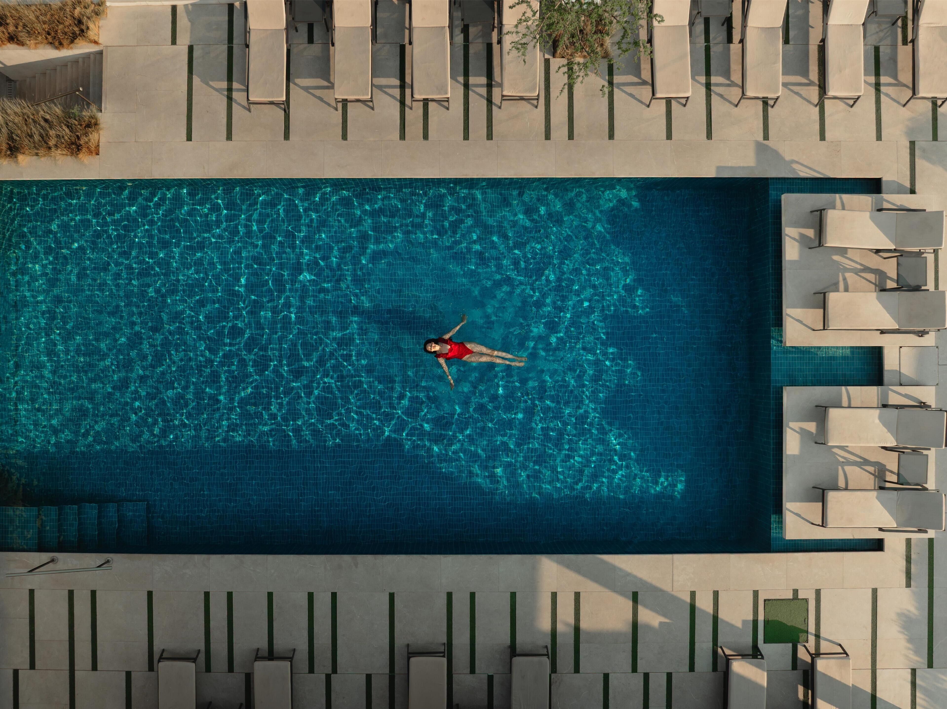 An aerial view of a rectangular swimming pool with a person floating in the center. Surrounding the pool are lounge chairs and a small garden area.
