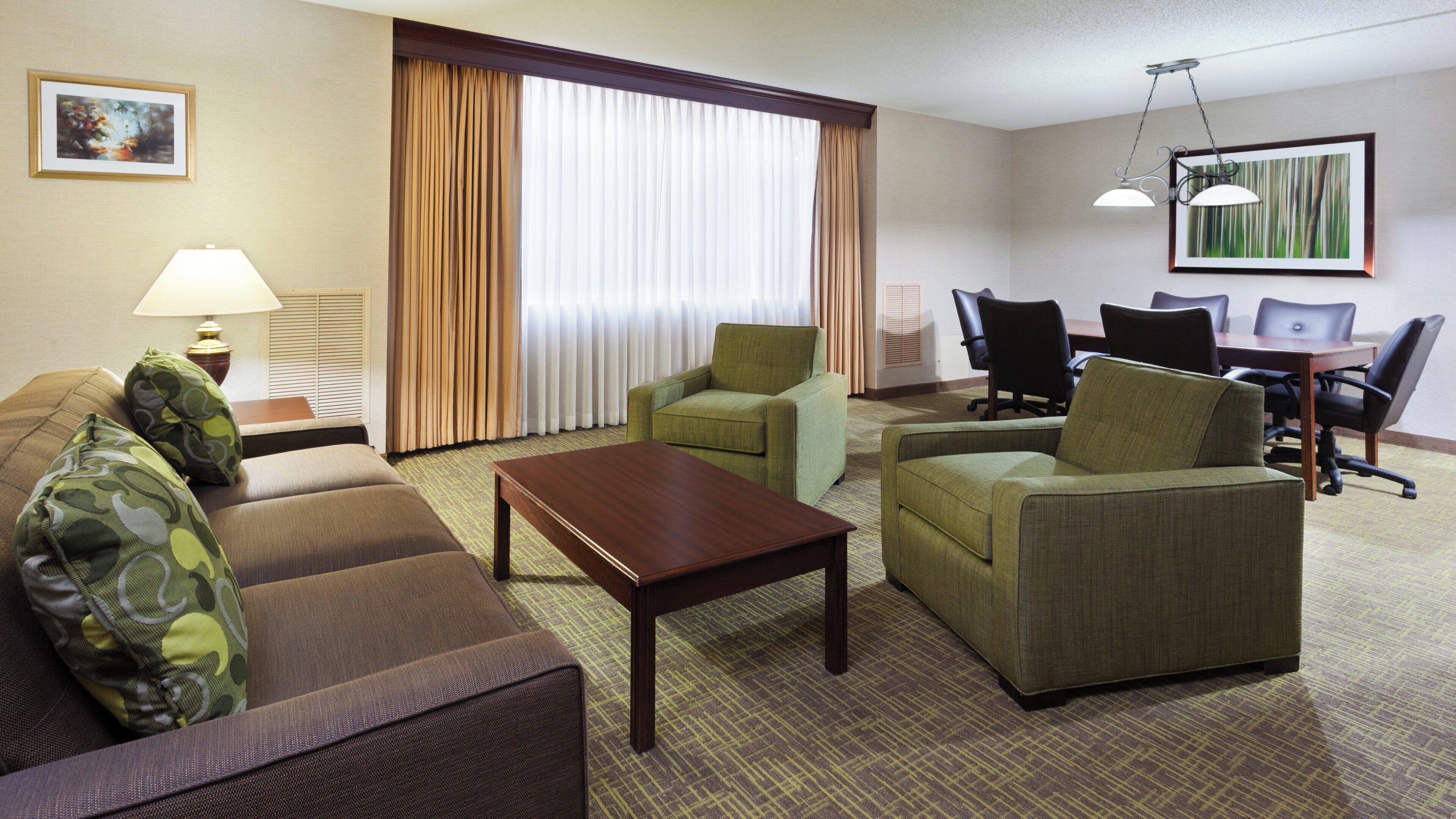 Habitación de hotel con sofá, sillones, una mesa de centro, una mesa de comedor con sillas, una lámpara y un cuadro en la pared.
