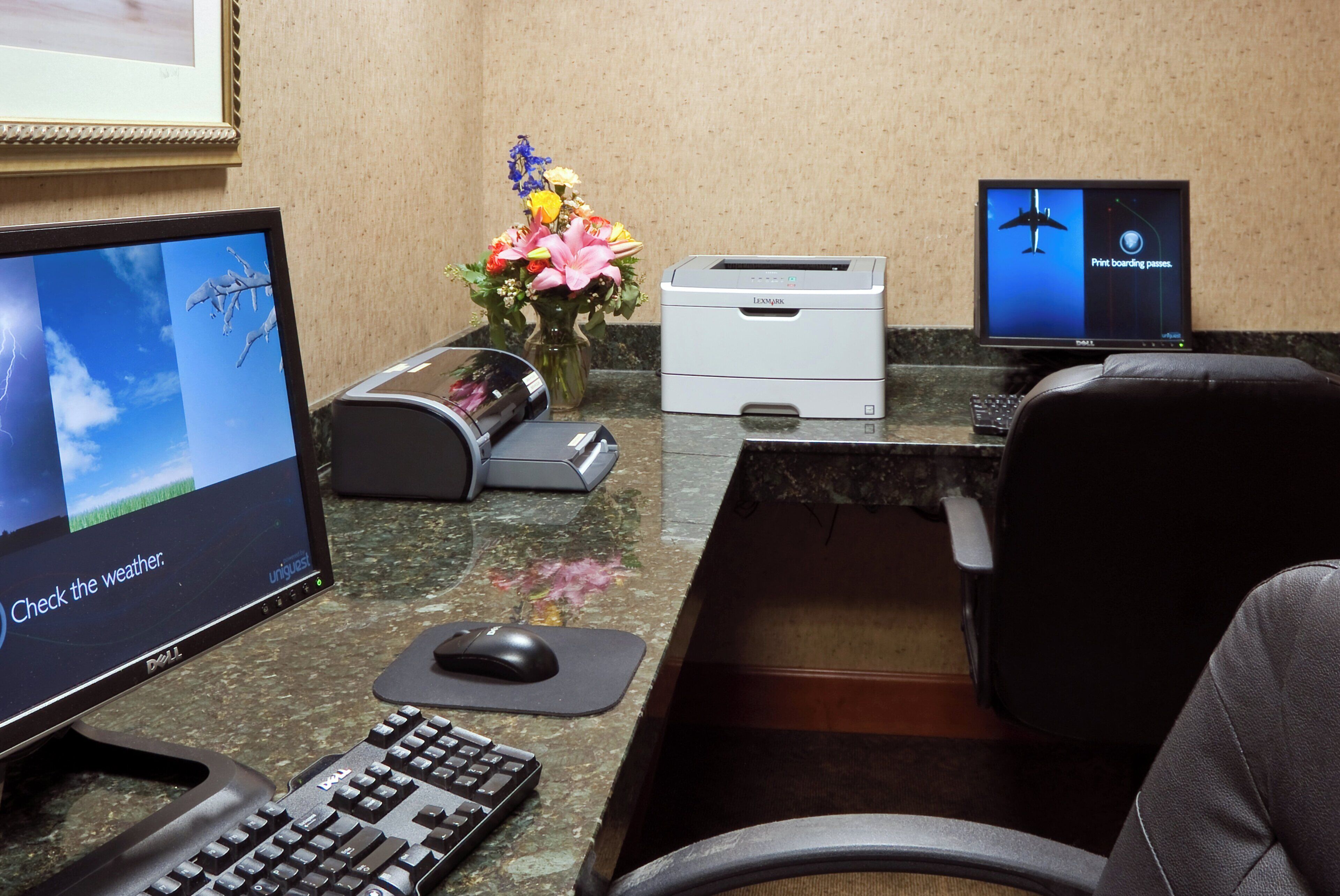 Un mostrador de recepción con un monitor de computadora que muestra un pronóstico del tiempo, una impresora, un teclado, un ratón y un florero con flores.