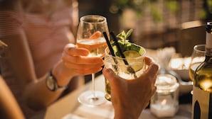 Una persona sosteniendo una copa de vino blanco con una pajita, otra copa con una rodaja de lima y menta, y una botella de vino blanco sobre una mesa.