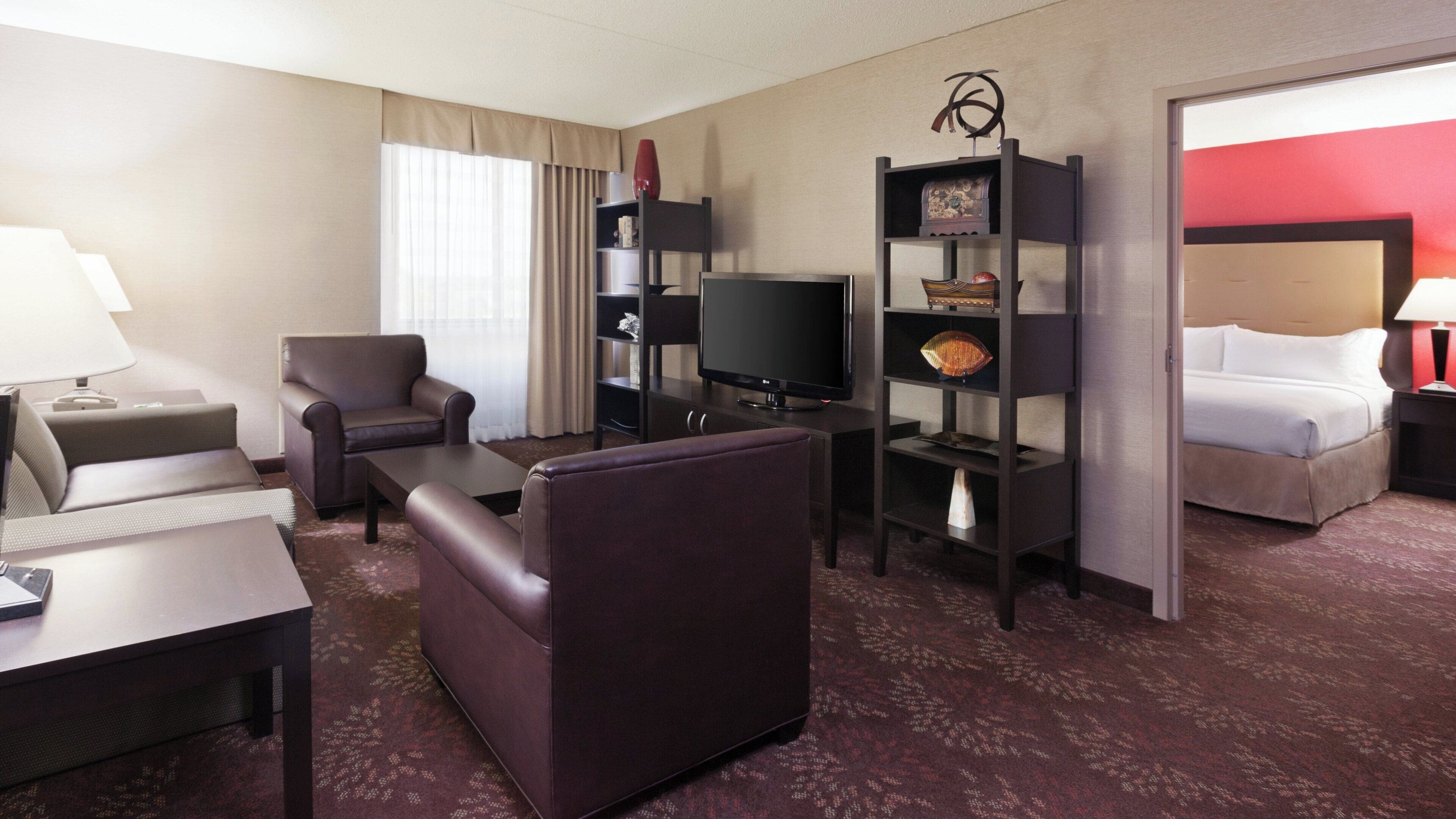 A hotel room with a sofa, armchair, TV, bookshelf, and a bed.