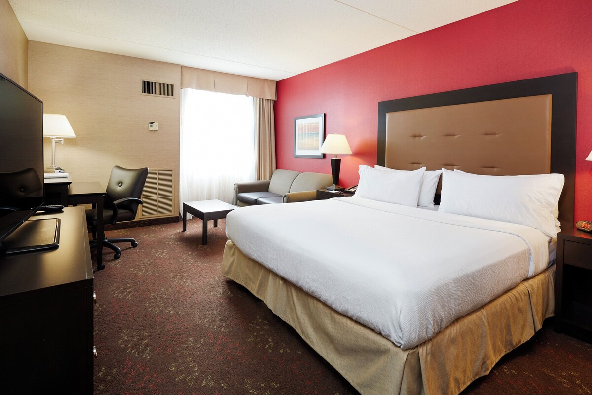 Une chambre d’hôtel avec un grand lit, un bureau avec un ordinateur, une chaise, une télévision et une fenêtre avec des rideaux.