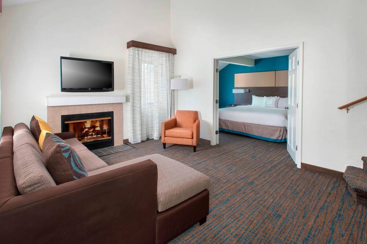 A hotel room with a sofa, a flat-screen TV mounted above a fireplace, a bed with a blue accent wall, and a door leading to another room.