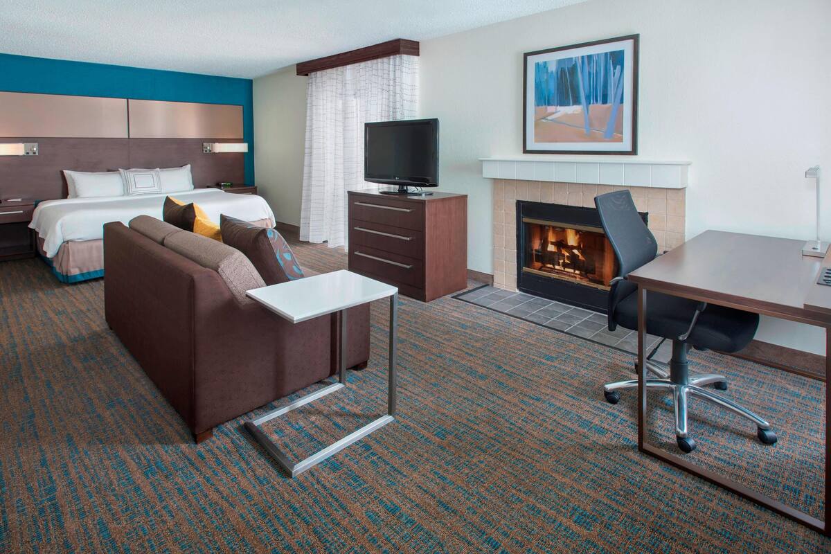 A hotel room with a bed, a sofa, a desk, a TV, and a fireplace.