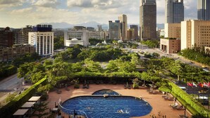 Hotel com piscina, rodeado por vegetação e edifícios modernos ao fundo.