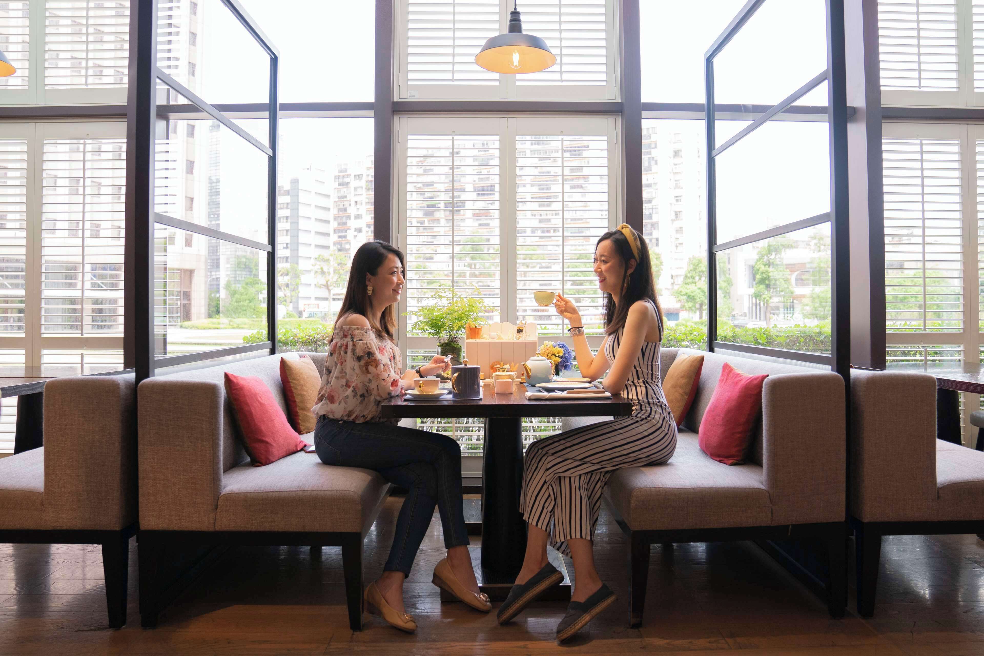 Duas pessoas sentadas à mesa numa divisão com grandes janelas, um sofá e um candeeiro pendente.