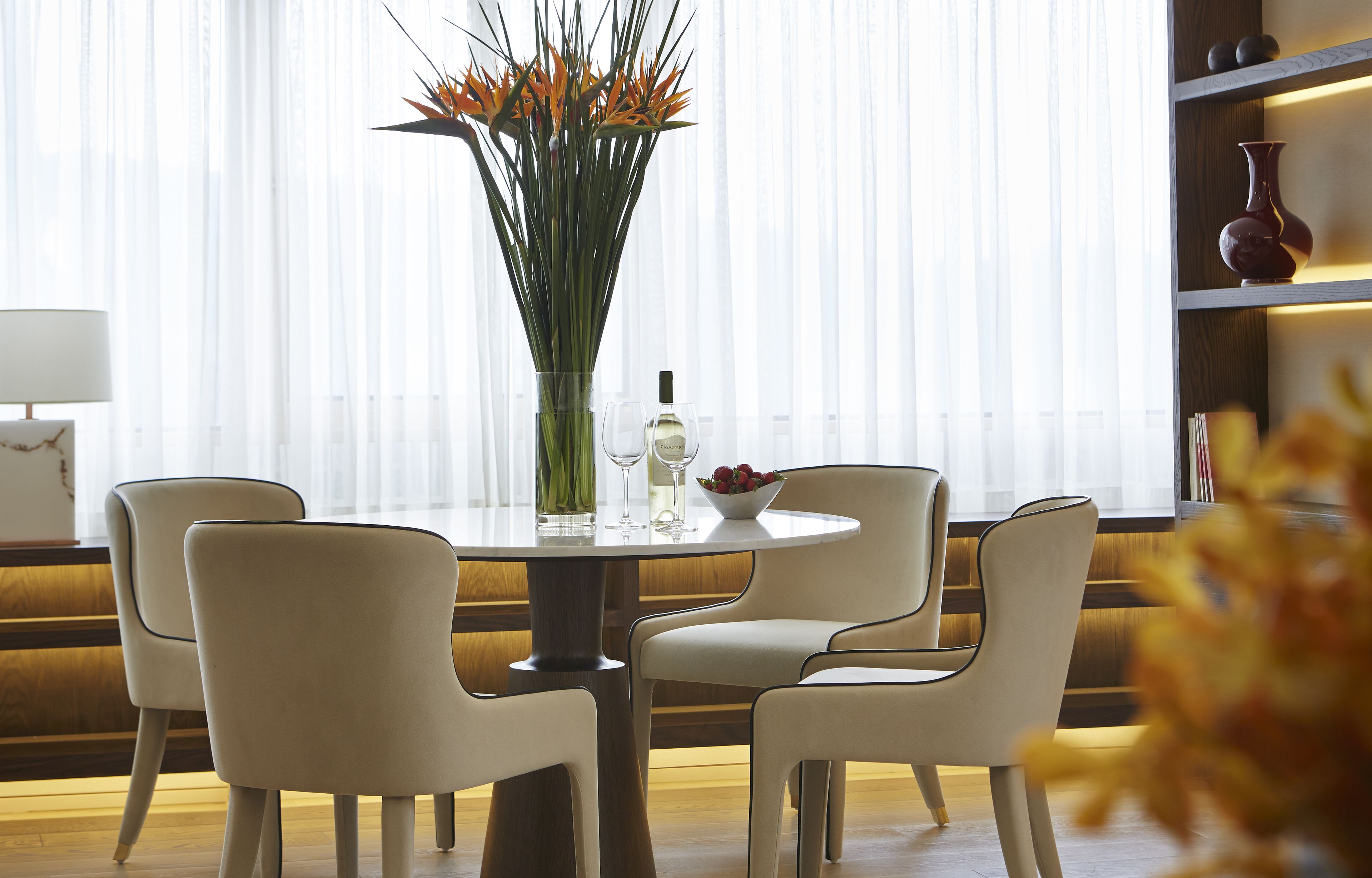 A dining area with a round table, four chairs, a vase with flowers, and a wine bottle.