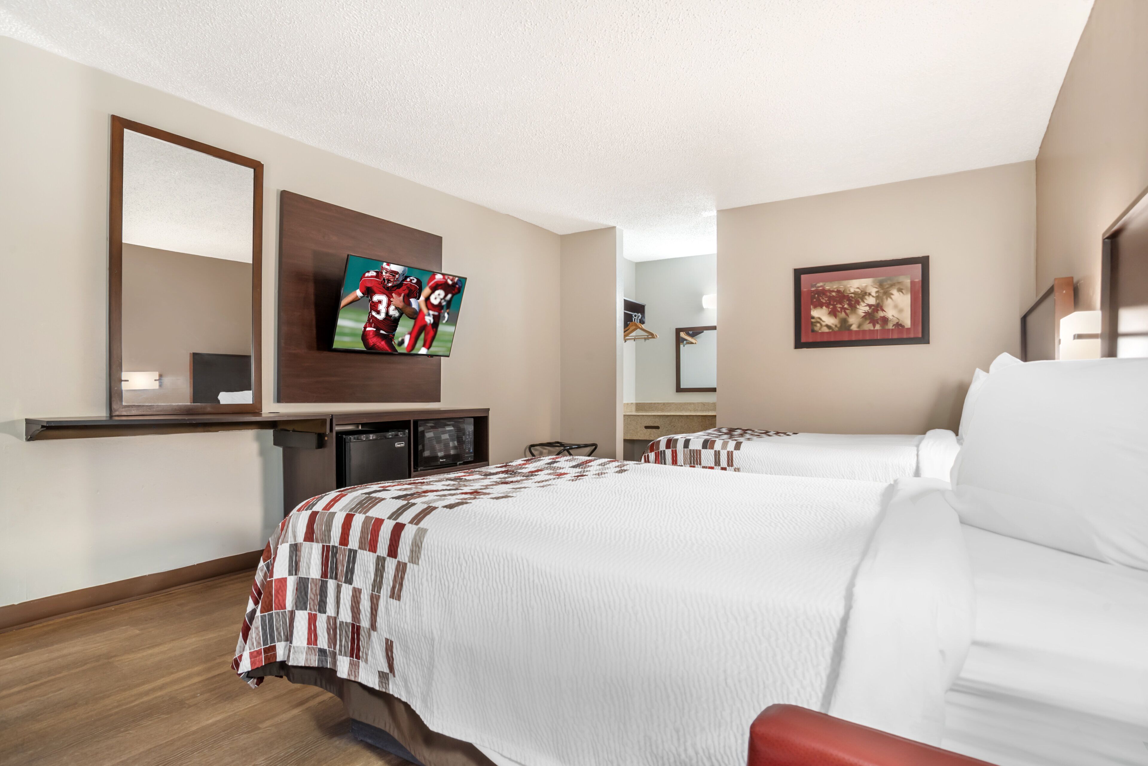 Ein Hotelzimmer mit zwei Betten, einem Flachbildfernseher, einem Spiegel, einem Schreibtisch mit einem Computer und einem gerahmten Bild an der Wand.