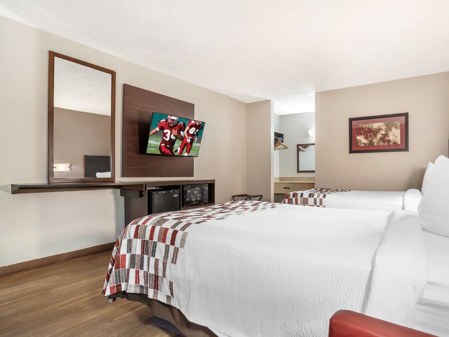 Ein Hotelzimmer mit zwei Betten, einem Flachbildfernseher, einem Spiegel, einem Schreibtisch mit einem Computer und einem gerahmten Bild an der Wand.