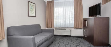 Ein Hotelzimmer mit grauem Sofa, einem Fernseher auf einem Holzschrank, einem Fenster mit Vorhängen und einem gemusterten Teppich.