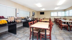 A cafeteria with multiple tables and chairs, a buffet counter with food items, and a television mounted on the wall.