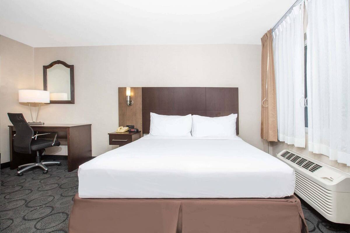Habitación de hotel con cama, escritorio con silla, teléfono, lámpara, espejo y ventana con cortinas.