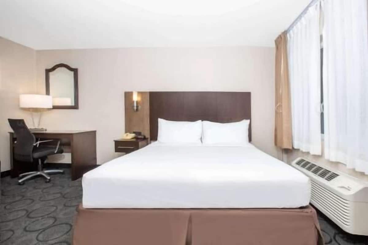 Habitación de hotel con cama, escritorio, silla, mesita de noche, lámpara, espejo y ventana con cortinas.