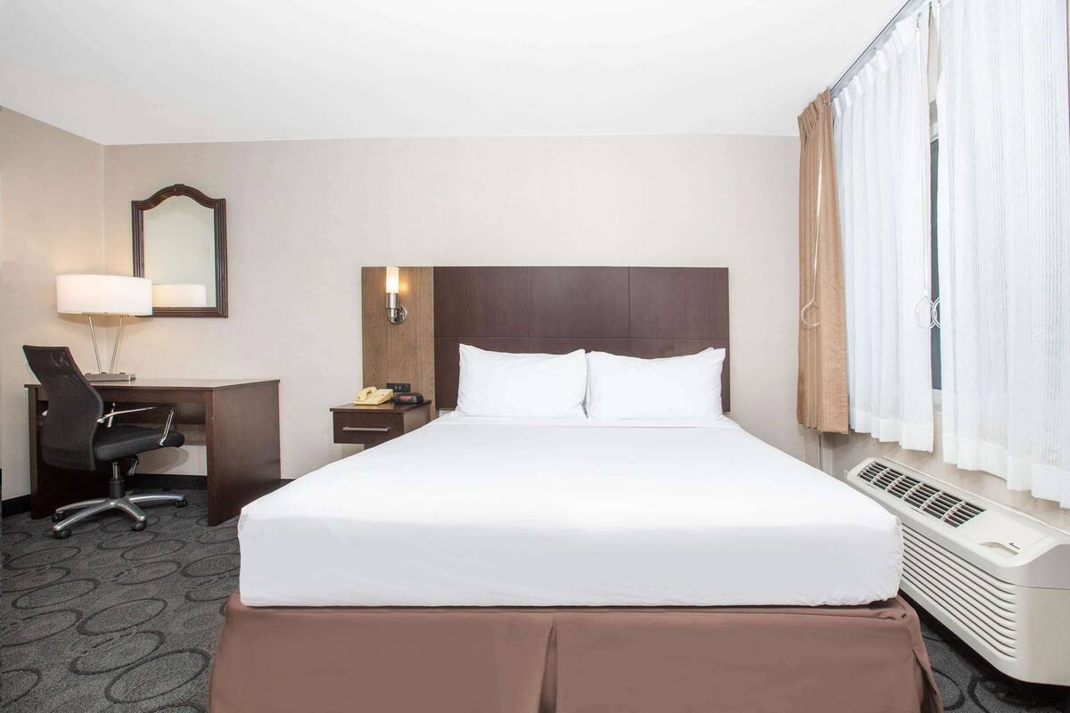 Habitación de hotel con cama, escritorio con silla, teléfono, espejo y ventana con cortinas.