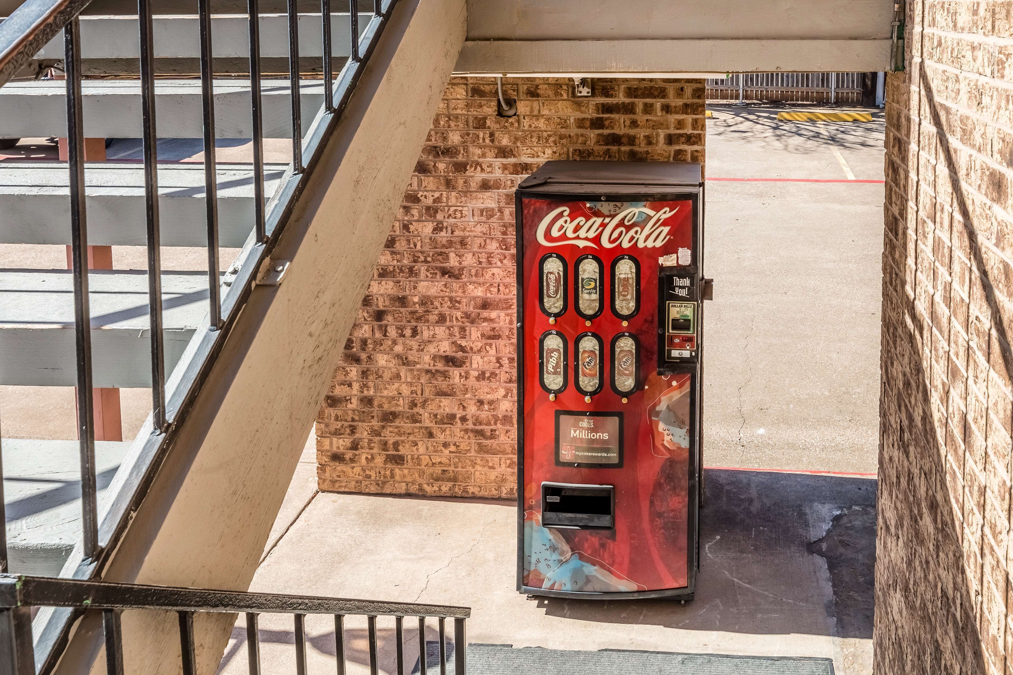 A Coca-Cola vending machine on a sidewalk next to a brick wall and a metal staircase.