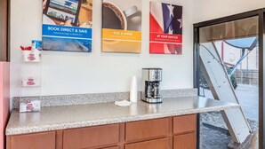Un espace d’accueil d’hôtel avec une machine à café, une cafetière en verre et un ensemble de tasses à café sur le comptoir. Trois affiches sont accrochées au mur, faisant la publicité de différents services.