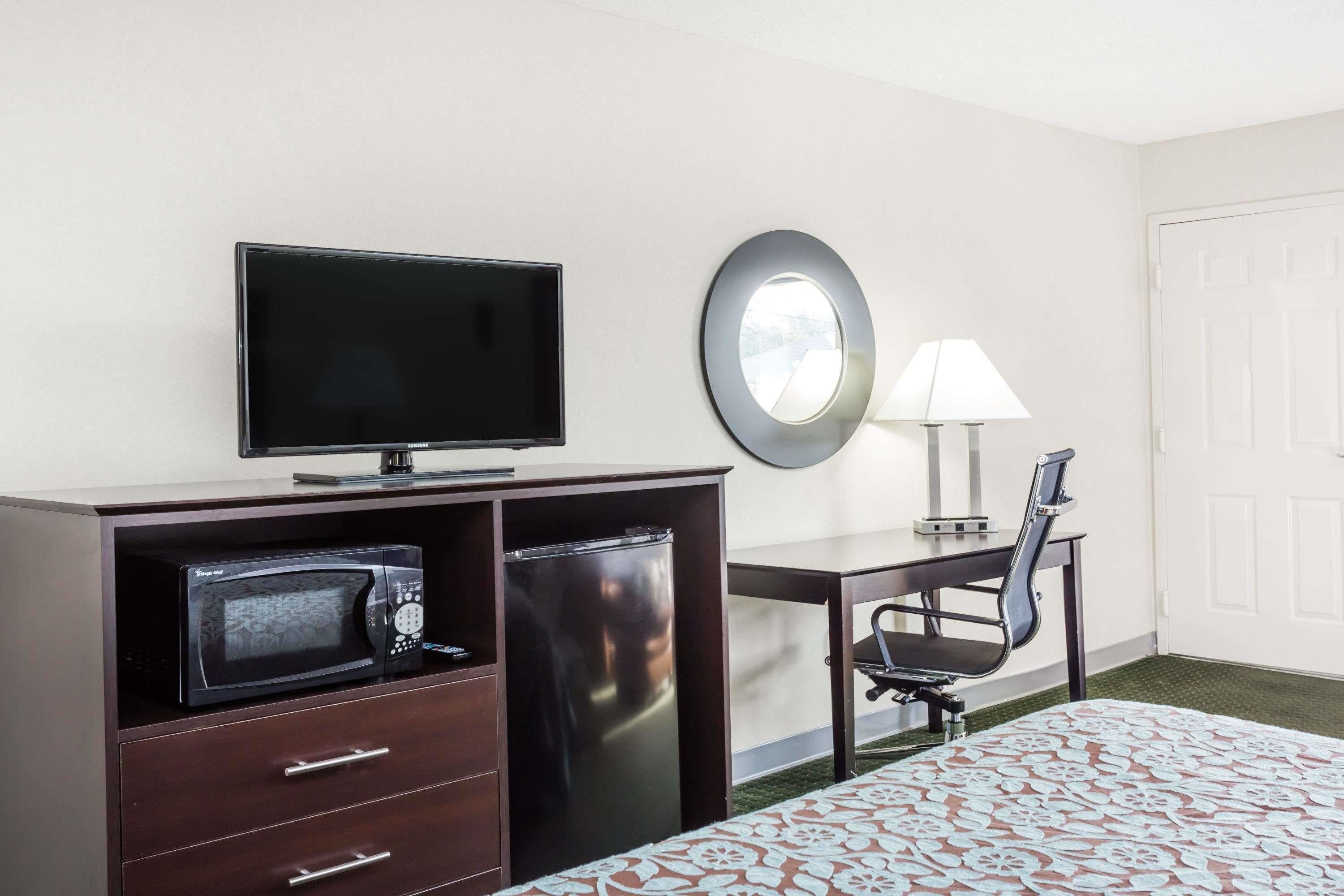 A hotel room with a bed, a desk with a chair, a television, a microwave, a refrigerator, a lamp, and a door.