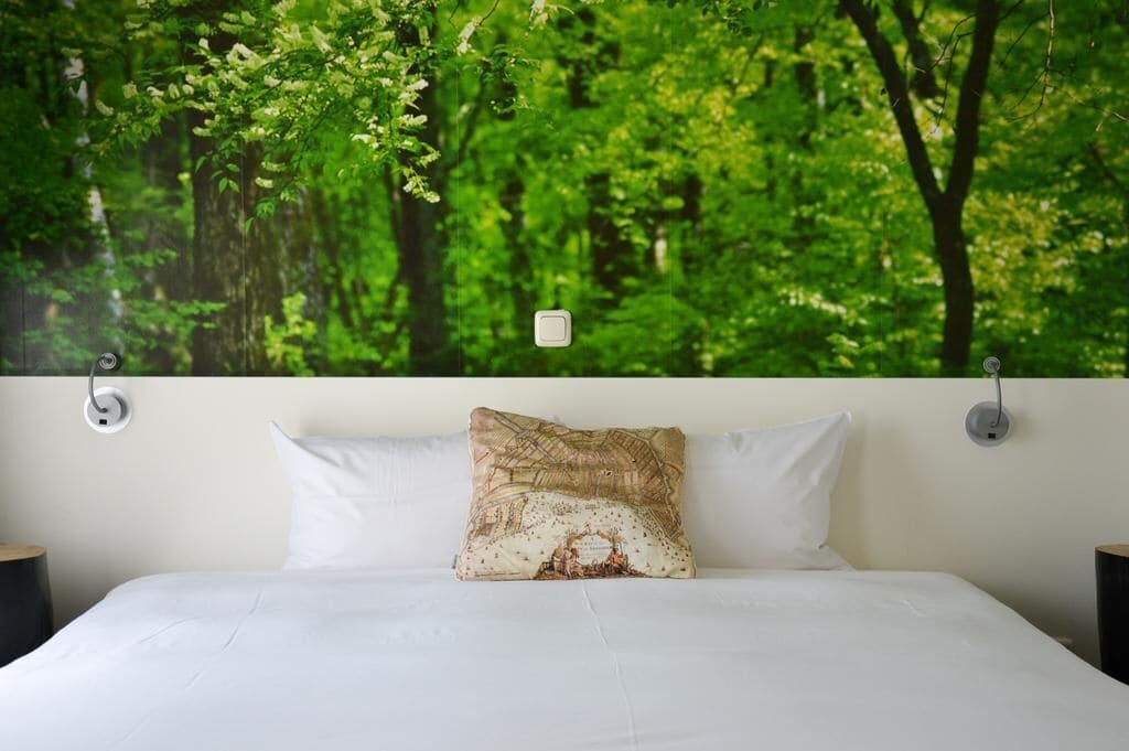 A bed with white linens, a patterned pillow, and a wall-mounted light fixture.