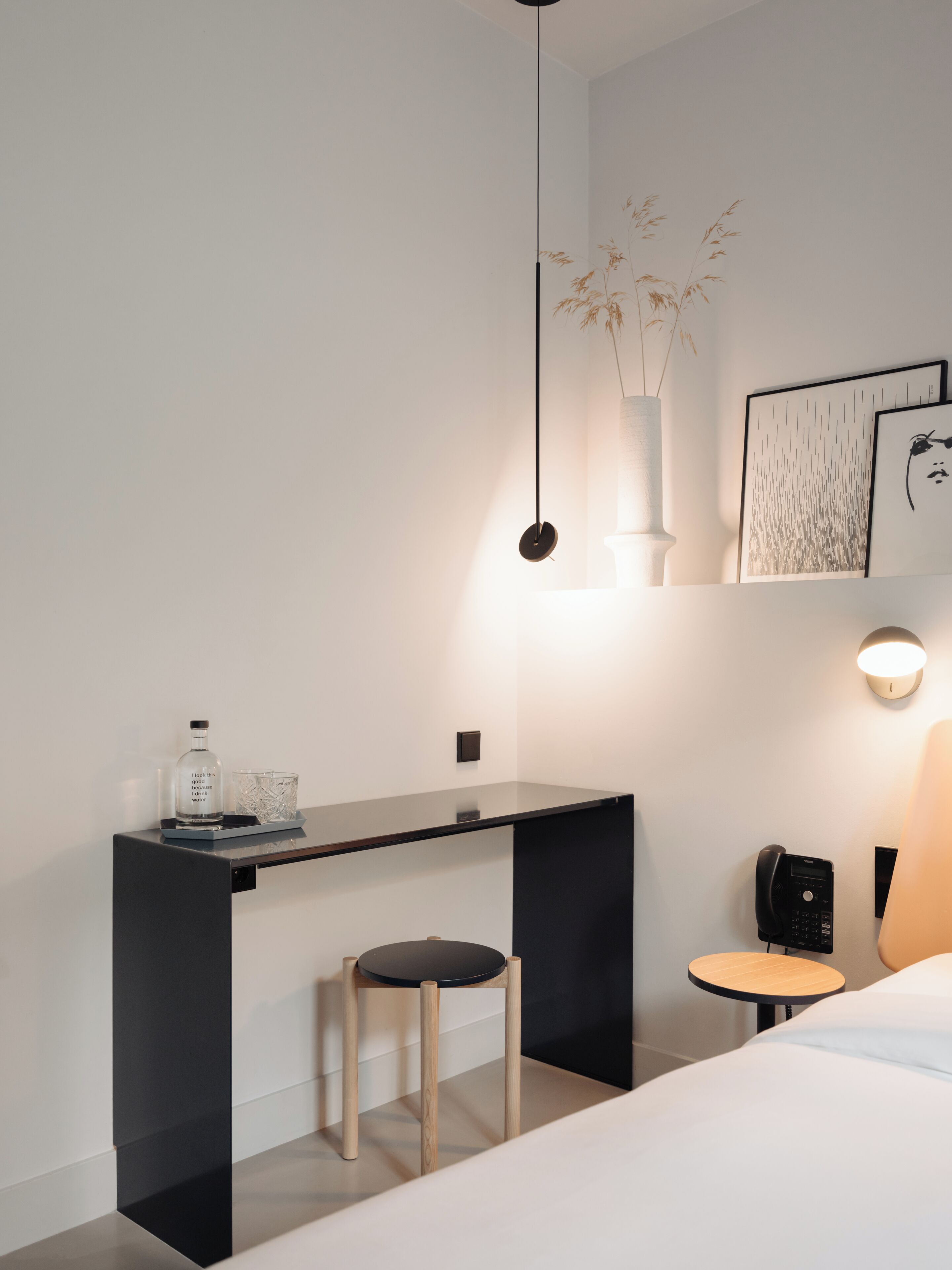 Une chambre d’hôtel moderne avec un bureau au design épuré, un petit tabouret, une table de chevet et un lit.