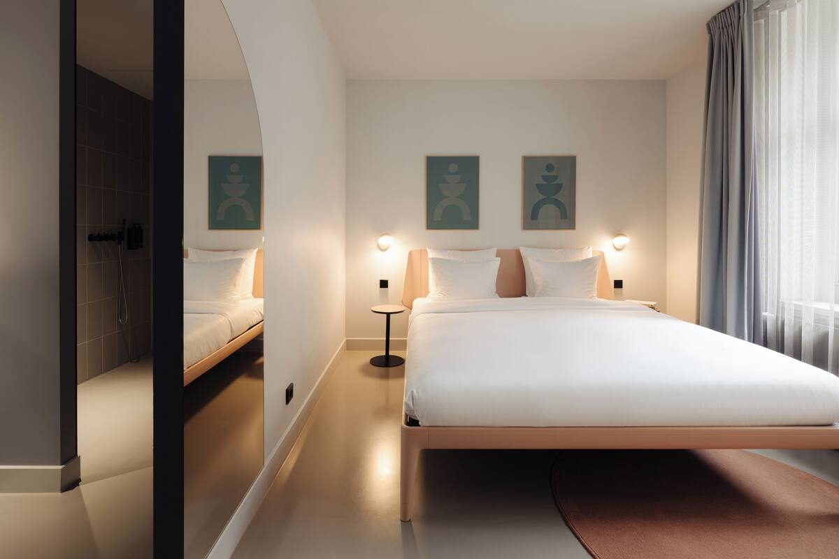 A modern hotel room with a large bed, a small side table, a wall-mounted light, and a shower area.