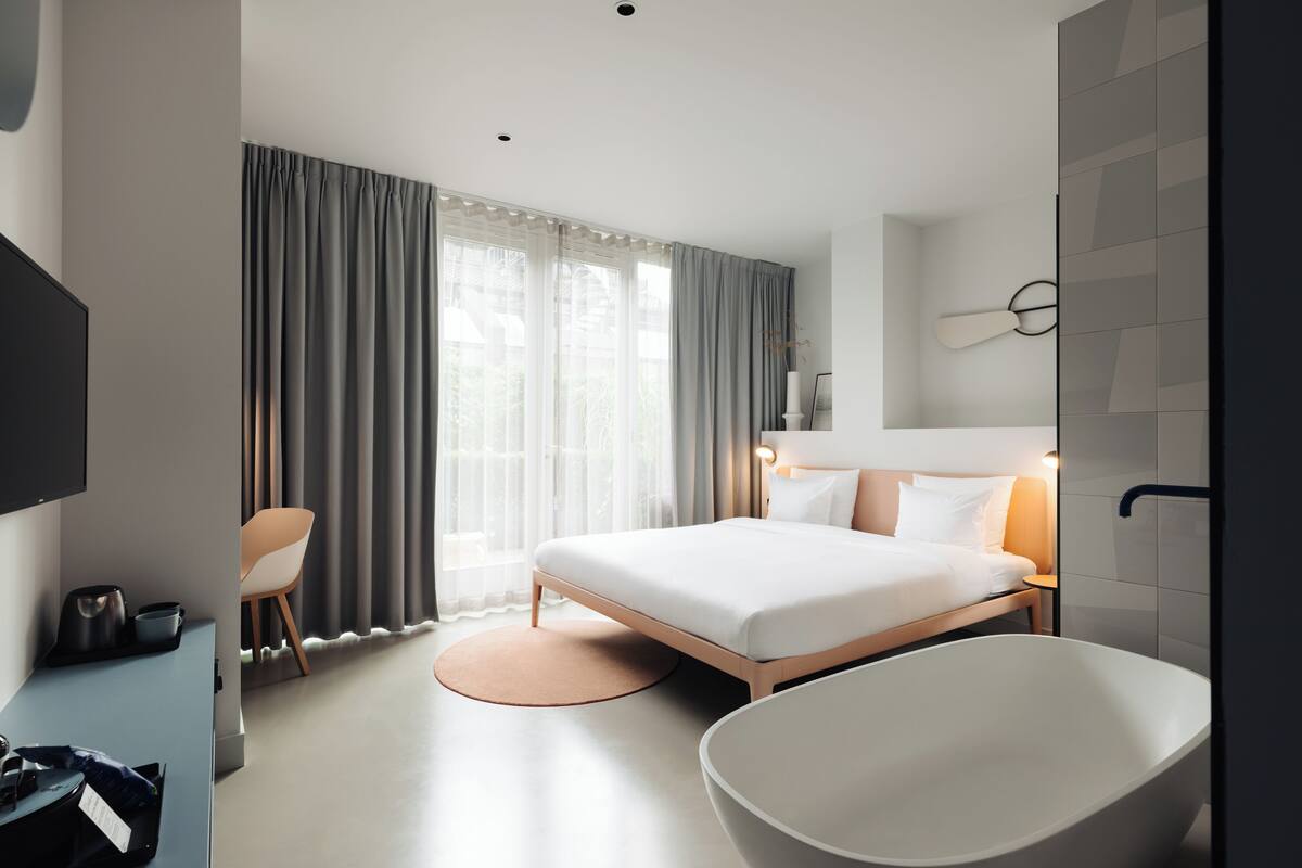 A modern hotel room with a large bed, a bathtub, a desk with a chair, and a TV.